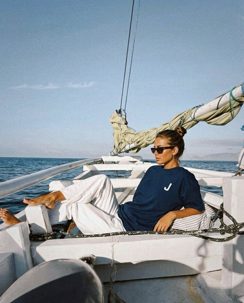 Person sitting on a sailboat with ocean and sky in the background wearing Baron & Buck navy shirt with embroidered letter "J"