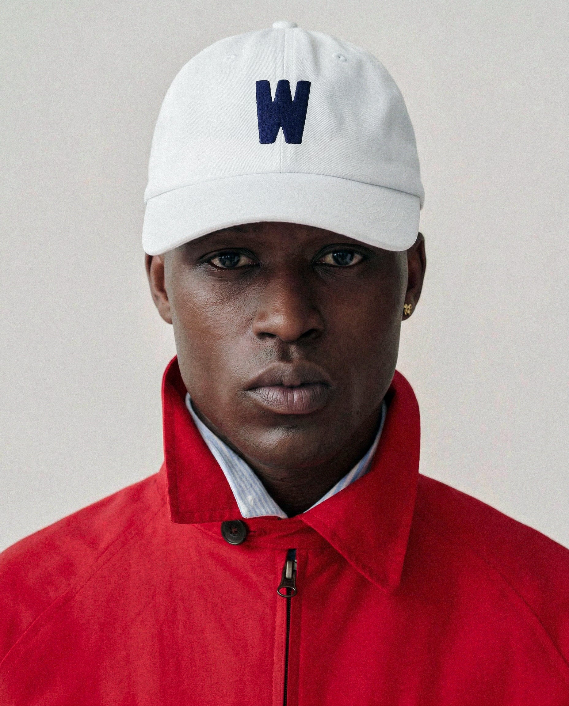 Person wearing  Baron & Buck  white cap with a blue 'W' embroidered letter and a red jacket on a plain background