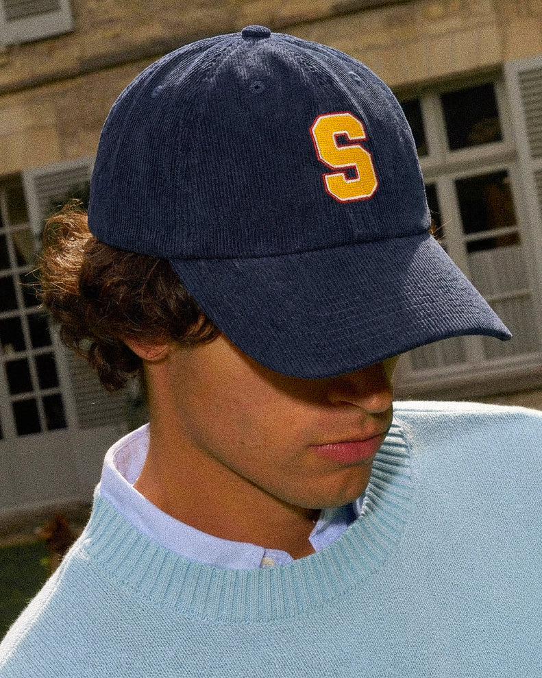 Person wearing a Baron & Buck corduroy navy blue cap with a yellow 'S' logo in front of a building.